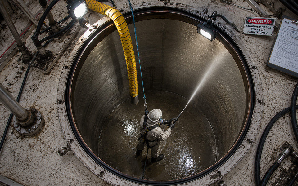 Worker performing confined space hydroblasting inside a large industrial tank with ventilation hose, safety harness, temporary work lighting, and confined space entry equipment during tank cleaning operations.