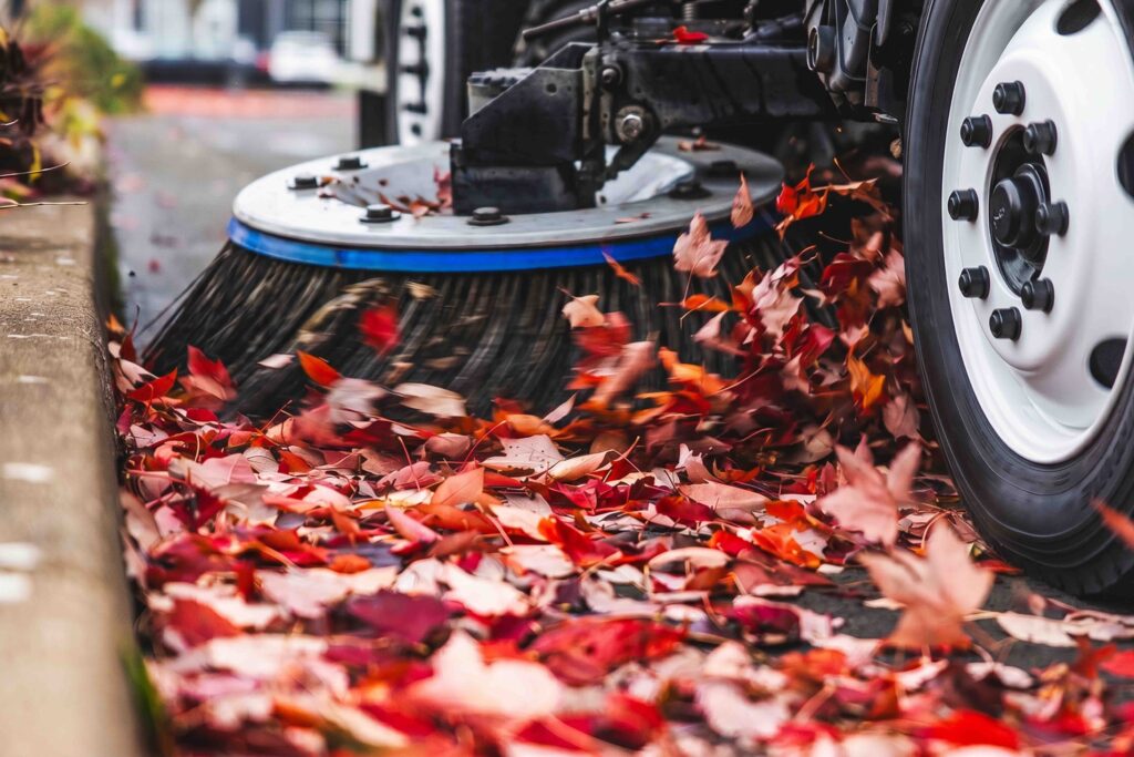 Street sweeper brush clearing red fall leaves from a curbside parking area.
