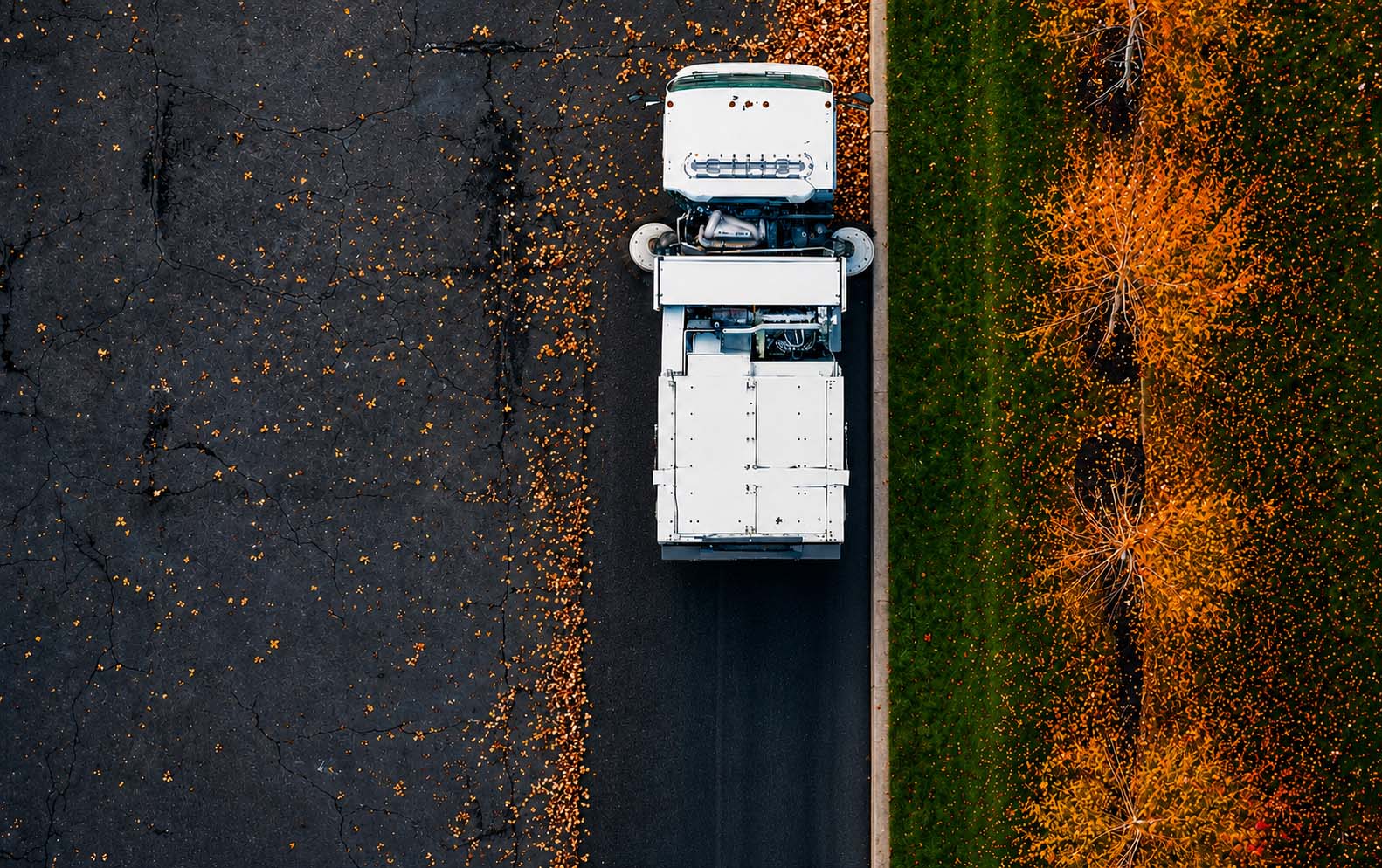 Aerial view of a street sweeper cleaning fall leaves along a curb beside a landscaped commercial property.