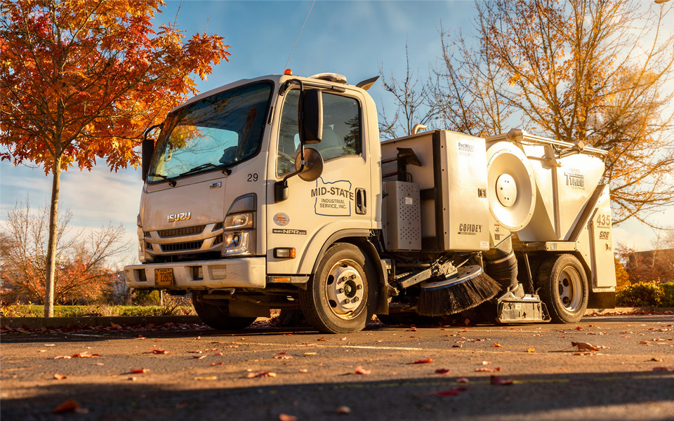 Mid-State Industrial Service Tymco air sweeper parked along a curb during fall leaf cleanup operations in a commercial parking lot.