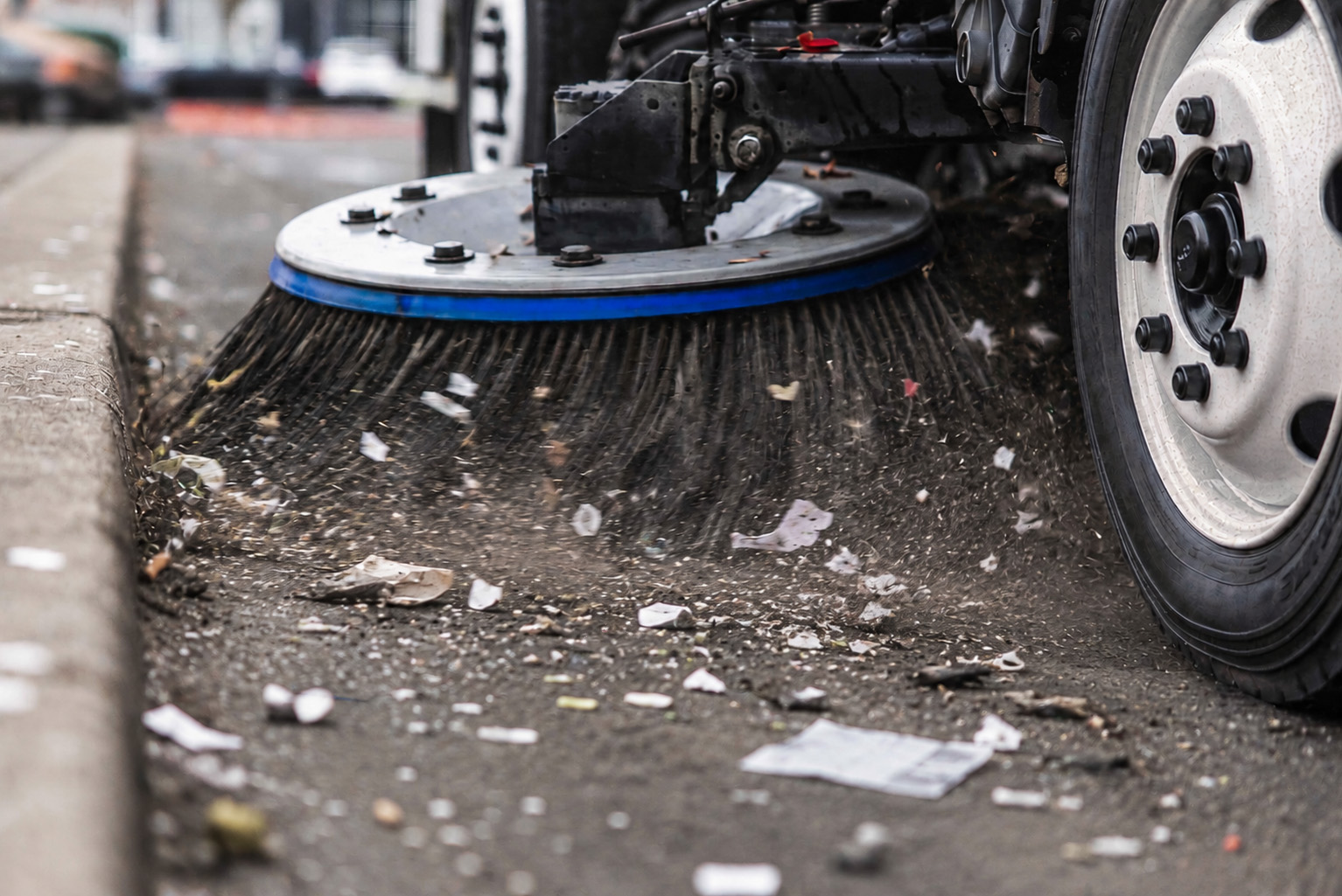 Street sweeper brush cleaning dirt, paper scraps, and small debris from a parking lot curb.