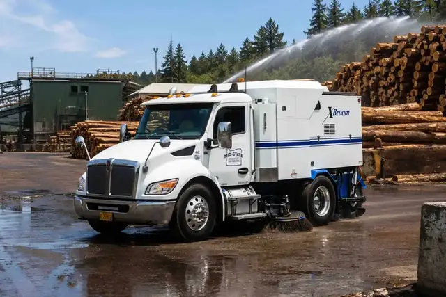 Mid-State sweeper cleaning wet pavement in a log yard with a sprinkler operating behind it.