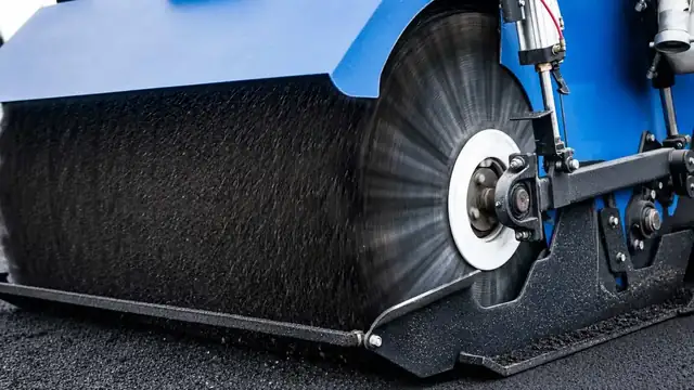 Close-up of a mechanical sweeper’s cylindrical brush clearing loose asphalt and aggregate from pavement.