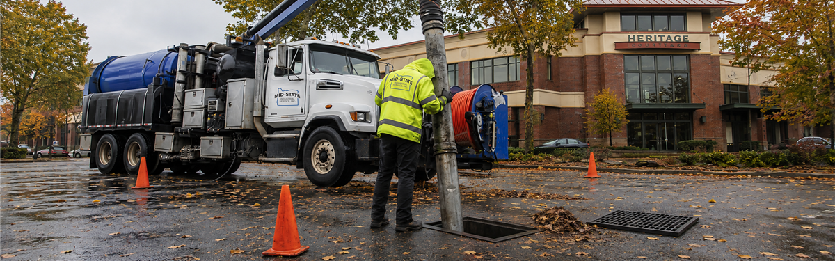 Mid-State worker vacuum-cleaning an open parking lot storm drain, with the removed grate and fallen leaves nearby.