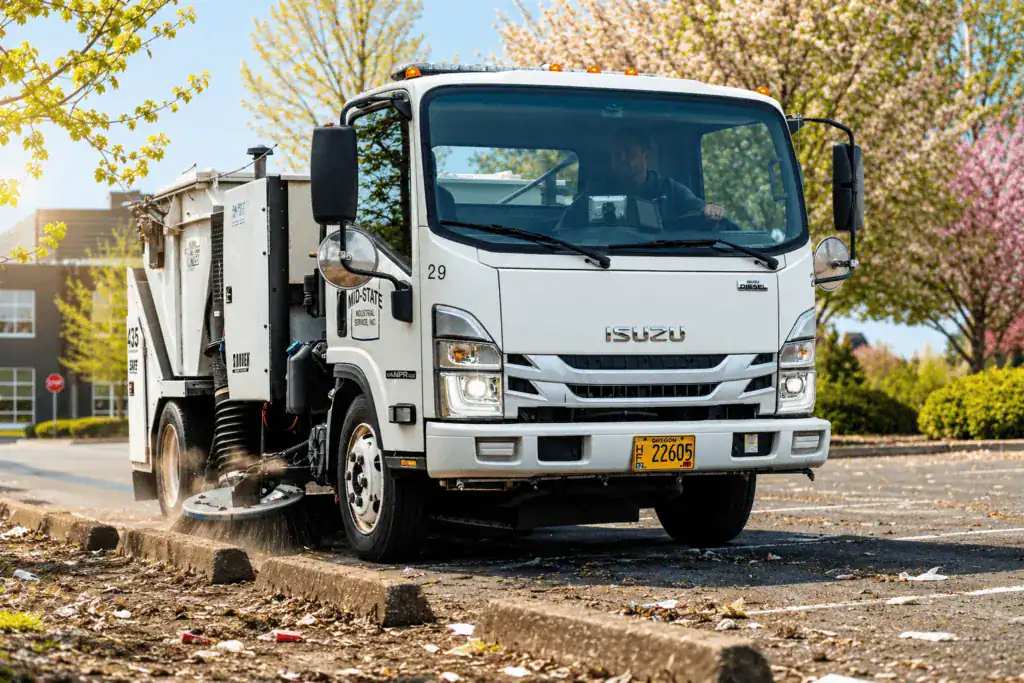 Mid-State TYMCO sweeper clearing leaves and debris along a parking lot curb.